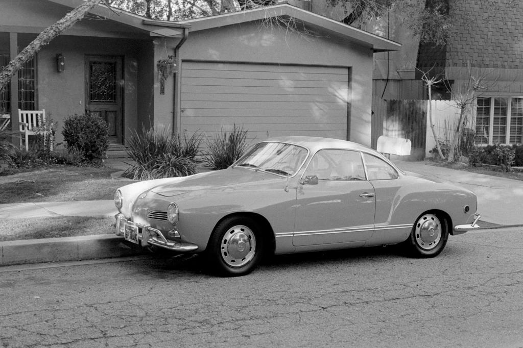 Black and white image of a Harmann Ghia coupe parked outside of a house.